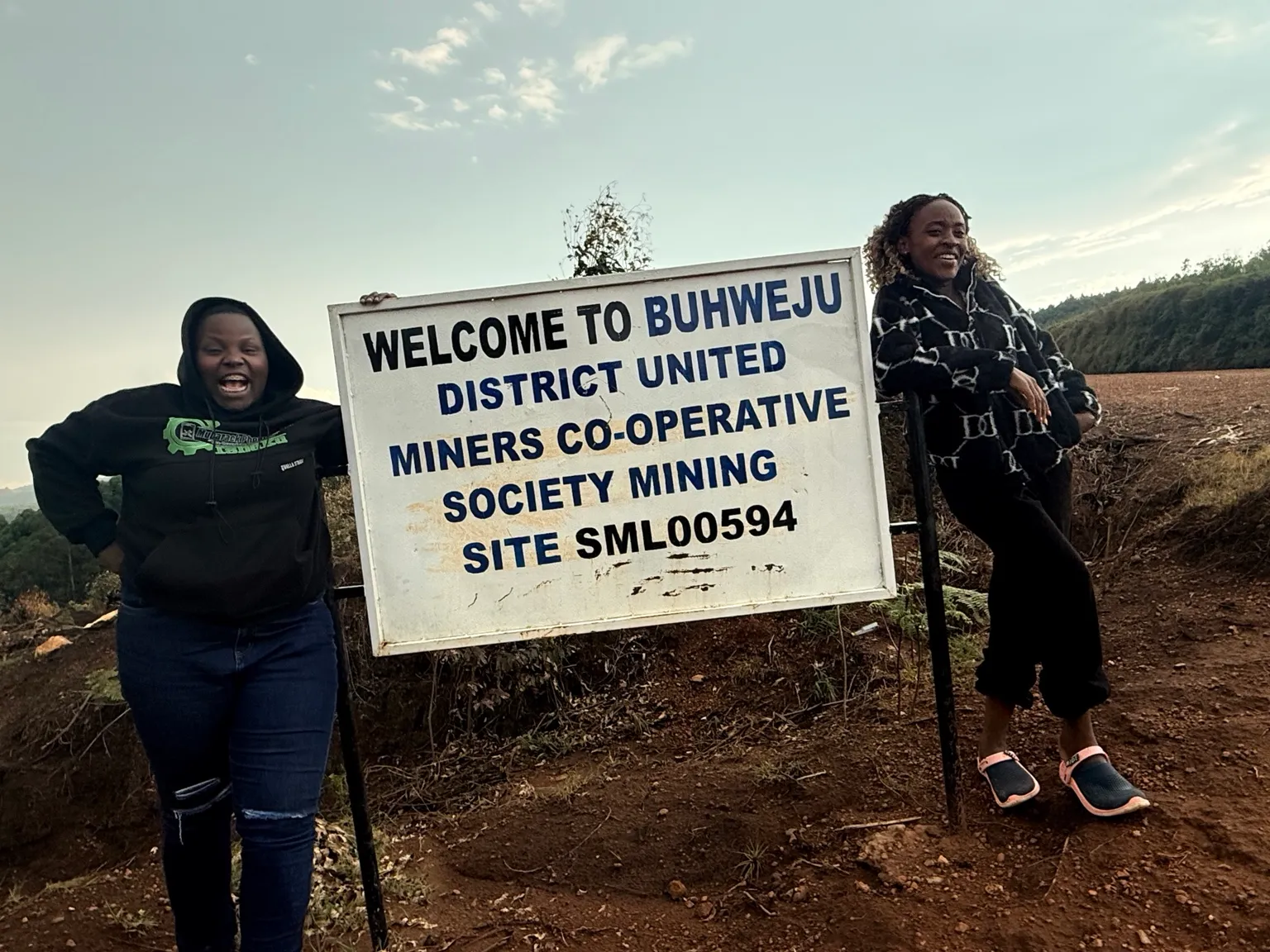 Two people standing either side of a painted sign reading Welcome to Buhweju District United Miners Co-operative Society Mining Site SML00594, on bare red earth with cleared ground behind
